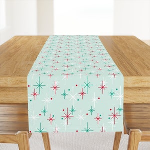 May include: A light blue table runner with a white, red, and green starburst pattern. The runner is on a wooden table.