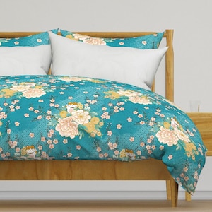 May include: A teal blue duvet cover with a floral pattern featuring white peonies, pink cherry blossoms, and gold accents. The pattern also includes small orange cats with gold accents. The duvet cover is on a wooden bed frame.