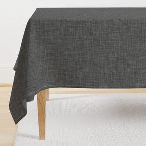 Puede incluir: Un mantel de color gris oscuro con un sutil patrón texturizado. El mantel está drapeado sobre una mesa con patas de madera.