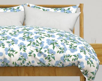 May include: White duvet cover with a blue floral pattern featuring hydrangeas. The duvet cover is on a wooden bed frame with a white pillow.