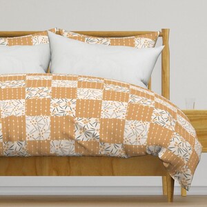 May include: A patchwork quilt and pillow set in a bedroom setting. The quilt features a repeating pattern of squares in shades of orange, cream, and gray. The design includes floral and dotted elements. The bed frame is made of light-colored wood.