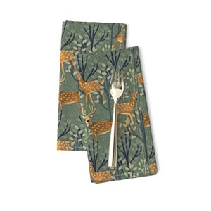 May include: Two green fabric napkins with a repeating pattern of brown deer in a forest setting. The deer have white spots and are surrounded by green leaves and branches.