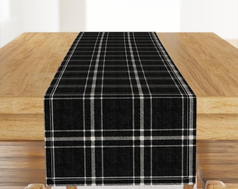 May include: A rectangular table runner featuring a black and white plaid pattern. The fabric displays a woven texture with intersecting thin white lines forming a grid over a solid black background, creating a classic tartan-inspired design.