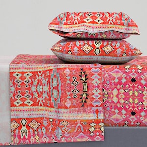 May include: Two decorative pillows and a table runner with a vibrant, geometric pattern in shades of red, pink, and blue. The pillows are stacked on top of the runner, creating a layered effect. The textile design features a mix of diamond and rectangular shapes.