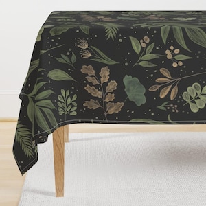 Op de afbeelding: Een zwarte tafelkleed met een botanisch patroon van groene en bruine bladeren, takjes en bessen. Het ontwerp bevat verschillende bladvormen en -groottes, met kleine witte spikkels. Het tafelkleed ligt over een houten tafel.