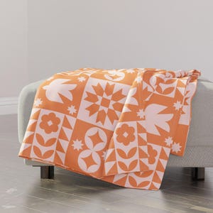 May include: A soft orange and white throw blanket with a geometric pattern. The blanket is folded and draped over a couch.