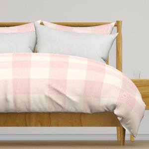 May include: A bed with a light wood frame and a duvet cover featuring a large pink and white gingham pattern. Two white pillows are visible, along with a wooden nightstand.