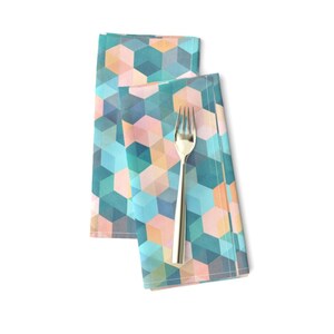 May include: Two folded napkins with a geometric pattern of teal, blue, pink, and peach squares. The napkins are folded in thirds and are lying on a white surface.