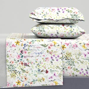 May include: A bedding set featuring two rectangular pillows stacked on top of a flat sheet. The fabric is white and decorated with a watercolor-style print of various small flowers and green leaves in shades of pink, yellow, purple, and green.