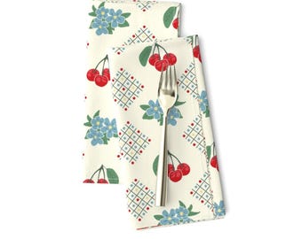 Op de afbeelding: Twee witte stoffen servetten met een herhalend patroon van rode kersen, blauwe bloemen en een geel en blauw geometrisch ontwerp.