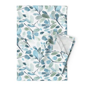 May include: A folded white rectangular cloth with a watercolor pattern of blue and green leaves and branches. The leaves vary in shades of blue and green, creating a soft, natural design. The cloth is folded over, revealing a white interior.