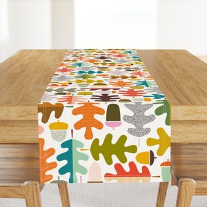 May include: A colorful table runner featuring a repeating pattern of stylized leaves and acorns in shades of orange, green, teal, and brown on a cream background. The runner is displayed on a wooden table.