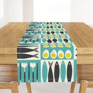 Op de afbeelding: Een turquoise en crèmekleurige tafelrunner met een geometrisch patroon van vissen, eieren en vorken. Het patroon wordt over de hele runner herhaald.
