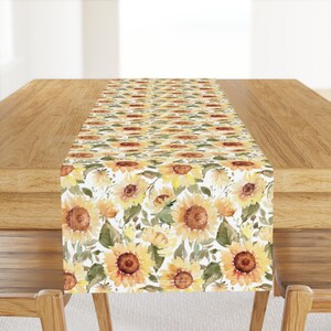 May include: A white table runner with a watercolor sunflower pattern. The sunflowers are yellow, orange, and brown with green leaves. The runner is on a wooden table.