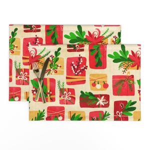 May include: A set of two beige placemats with a festive Christmas design. The placemats feature a repeating pattern of red and gold gift boxes with green holly and red berries.