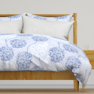 May include: A light blue duvet cover with a white background and a floral pattern of blue hydrangeas. The duvet cover is on a bed with a wooden headboard and two white pillows.
