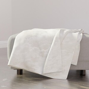 May include: A soft, white throw blanket with a subtle, cloudy, or marble-like pattern draped over a gray upholstered ottoman. The fabric features light gray, wispy, and ethereal textures that create a soft, layered appearance across the surface.