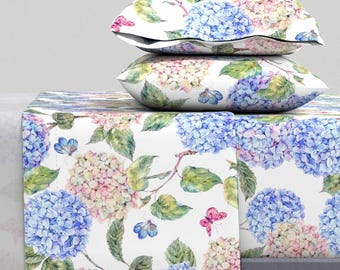 Puede incluir: Un juego de cama blanco con un estampado floral con hortensias y mariposas. El juego incluye una sábana plana, una sábana bajera y dos fundas de almohada.