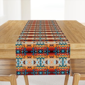 May include: A geometric patterned table runner in shades of orange, red, blue, and brown. The runner is made of fabric and has a fringed edge.