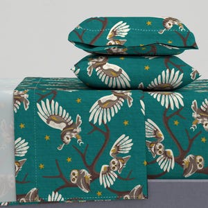 May include: Teal blue bedding set with a pattern of owls flying in front of branches with gold stars. The set includes a flat sheet, fitted sheet, and two pillowcases.