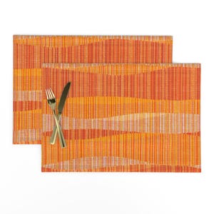 May include: Two orange and yellow placemats with a wavy striped pattern. A gold fork and knife are crossed on the placemat. The placemats are rectangular and made of a woven material. The colour scheme is warm and inviting.