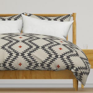以下が含まれることがあります： 明るい木製のフレームと模様入りの掛け布団カバーが付いたベッド。掛け布団カバーは、黒とグレーの幾何学模様で、小さな赤い十字のアクセントが付いています。白い枕が寝具セットを完成させています。