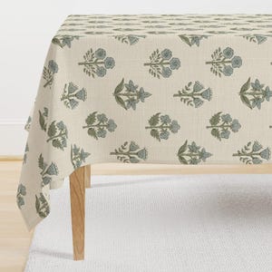 May include: A rectangular tablecloth with a cream-colored base featuring a repeating pattern of small, stylized floral sprigs in muted shades of sage green and dusty blue. The fabric has a visible woven texture and drapes over a wooden table.