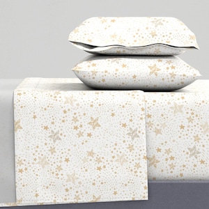 Puede incluir: Una sábana de cama blanca con un patrón de estrellas doradas. La sábana está doblada y apilada sobre dos fundas de almohada a juego.