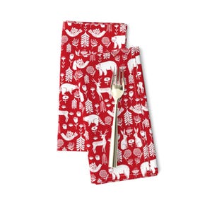 May include: Two red cloth napkins with a white Scandinavian-style pattern of animals, trees, and flowers. The napkins are folded and a silver fork is placed on top.