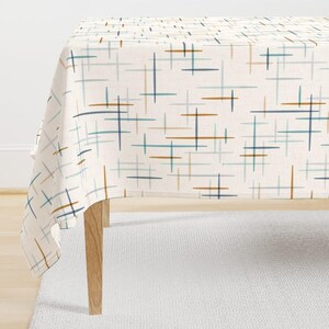 May include: A rectangular tablecloth featuring an off-white base with a repeating pattern of thin, horizontal and vertical lines in shades of dark blue, light blue, and tan. The fabric drapes over a wooden table with four visible legs.