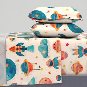 Op de afbeelding: Een wit beddengoedset met een ruimtethema. Het beddengoed heeft een raket, planeten, sterren en astronauten. De set bevat een hoeslaken, een plat laken en twee kussenslopen.