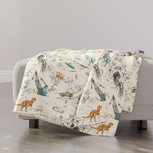 May include: Cream-coloured throw blanket with a whimsical design of dinosaurs, construction cranes, cars, and other vehicles. The illustrations are in shades of brown, green, and blue, creating a playful and imaginative pattern.