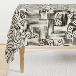 May include: A rectangular tablecloth with a cream background and a repeating black abstract pattern of curved and straight lines. The tablecloth is draped over a wooden table, with the legs visible.