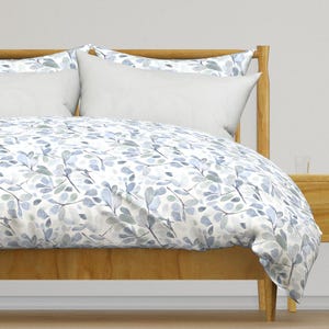 Puede incluir: Una funda nórdica blanca con un estampado de hojas de acuarela azul y verde. La funda nórdica está en una cama con una cabecera de madera y almohadas blancas.