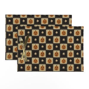 May include: A rectangular placemat featuring a repeating checkerboard pattern of black and beige squares. The beige squares contain orange jack-o'-lantern illustrations, while the black squares feature white stars. A silver fork and knife rest on top.