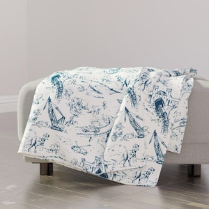 May include: A white throw blanket featuring a repeating toile-style pattern in dark blue ink. The design depicts various illustrations including construction cranes, dinosaurs, castles, helicopters, and cars scattered across the fabric surface.