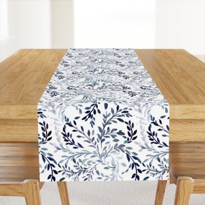 Pode incluir: Um corredor de mesa branco com um padrão floral azul e cinza. O corredor é feito de tecido e tem um brilho sutil.