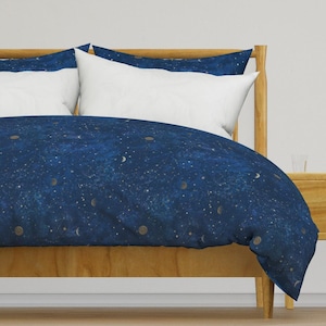 Op de afbeelding: Een donkerblauwe dekbedovertrek met een gouden sterrenhemelpatroon van sterren, manen en planeten. Het dekbedovertrek ligt op een houten bedframe met witte kussenslopen.