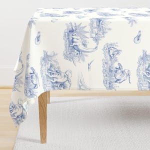 May include: A white tablecloth with a blue toile dinosaur pattern. The design features various dinosaurs and prehistoric plants in a classic toile style. The tablecloth is draped over a wooden table.