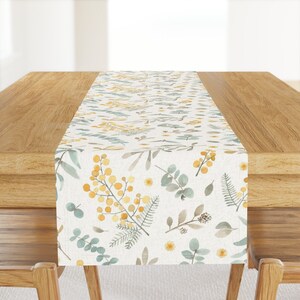 Könnte beinhalten: Ein weißer Tischläufer mit einem floralen Muster aus gelben und grünen Aquarellblumen und Blättern. Der Läufer liegt auf einem Holztisch.