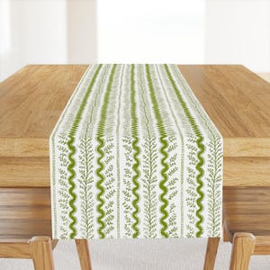 May include: A white table runner with a green leafy pattern. The pattern features repeating vertical lines of leaves and wavy lines.