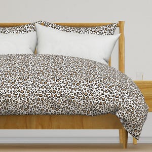 May include: A duvet cover and two pillow shams featuring a repeating leopard print pattern in shades of tan, brown, and black on a white background. The bedding set is displayed on a wooden bed frame with a headboard and a footboard.