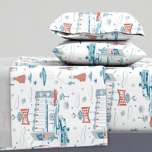 May include: White bed sheet set with a retro motel print in red, blue, and gray. The pattern includes cars, motels, palm trees, and figures. Two matching pillowcases are stacked on top of the sheet.