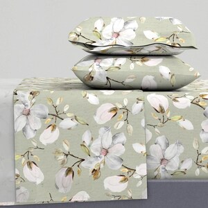 May include: A bedding set featuring two rectangular pillows stacked atop a flat sheet, all decorated with a repeating pattern of white magnolia flowers, brown branches, and green leaves on a light sage green background.