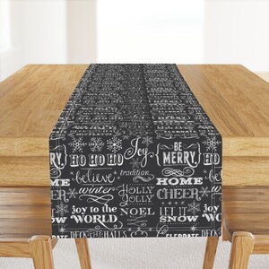May include: Black and white table runner with a chalkboard-style design featuring Christmas-themed words and snowflakes. The words include "Joy", "Be Merry", "Home Cheer", "Let It Snow", and "Deck the Halls".