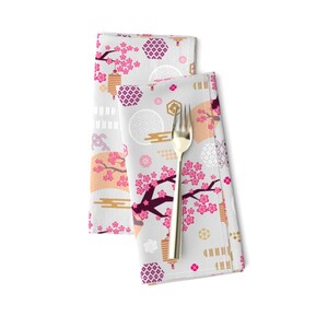 May include: Two white napkins with a pink and gold Japanese-inspired pattern. The pattern features cherry blossoms, lanterns, and geometric shapes. The napkins are folded and placed on a white surface.