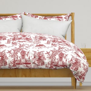 May include: A bed with a red and white toile duvet cover and pillowcases. The duvet cover features a detailed pastoral scene with figures, trees, and architectural elements. The bed frame is made of light-colored wood, and the pillows are white.