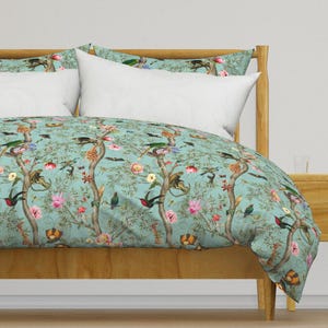 Op de afbeelding: Een tealblauwe dekbedovertrek met een bloemen- en dierenprint. De print bevat kolibries, vlinders en apen die in bomen klimmen. De dekbedovertrek ligt op een houten bedframe.