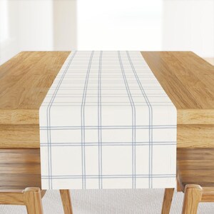 May include: A white table runner with a blue and white checked pattern. The runner is laid on a wooden table.
