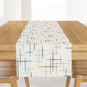 May include: A rectangular table runner featuring an off-white base fabric with a repeating pattern of thin, intersecting horizontal and vertical lines in shades of dark blue, light blue, and golden brown, creating a grid-like geometric design.